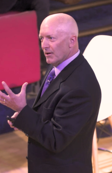 photo of a man in a dark suit and purple tie standing in front of a conference with an intense look on his face