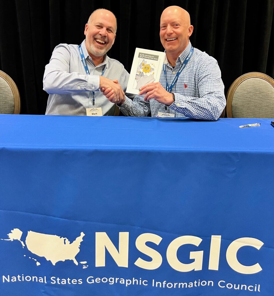 Two men sitting on a conference stage shaking hands and one of them is holding up the book he wrote