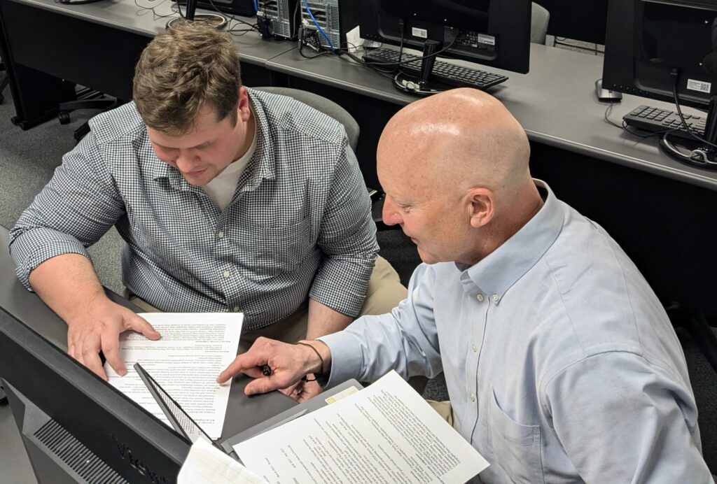 two men sit next to each other at a table, looking at documents and having a conversation.